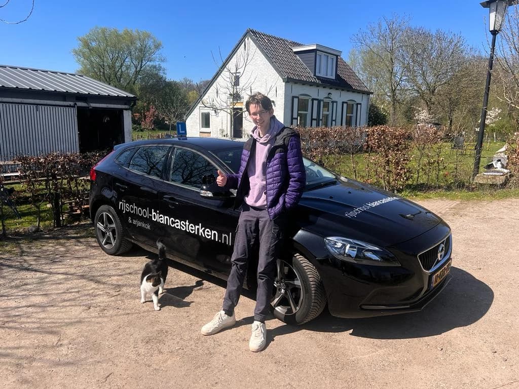 Tevreden leerling met duim omhoog naast de zwarte Volvo lesauto.
