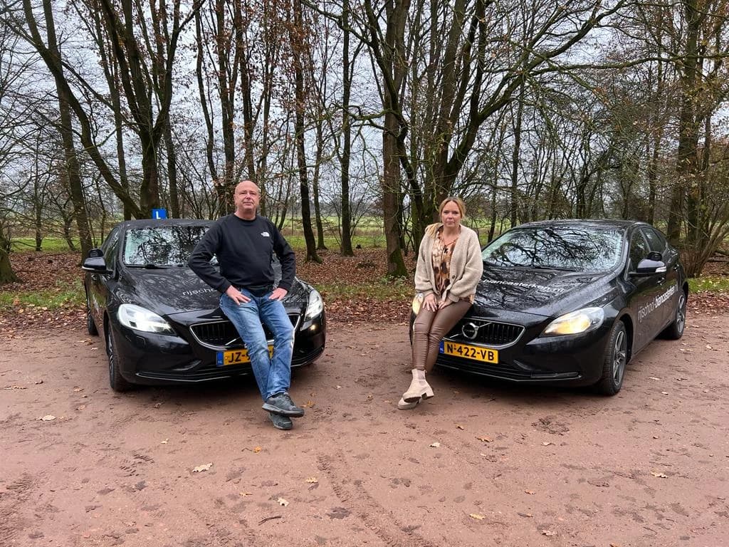 Rijinstructeurs Arjan Kok en Bianca Sterken zittend op de motorkap van hun lesauto's in het bos.
